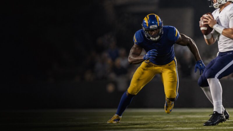 An edge rusher in blue and gold bends the corner towards the quarterback.