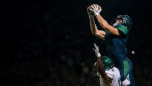 A receiver in navy and green reaches above a defender for the ball.